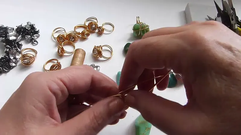 Manos que retuercen el alambre fino en la base del anillo, con una gema verde sobre la mesa