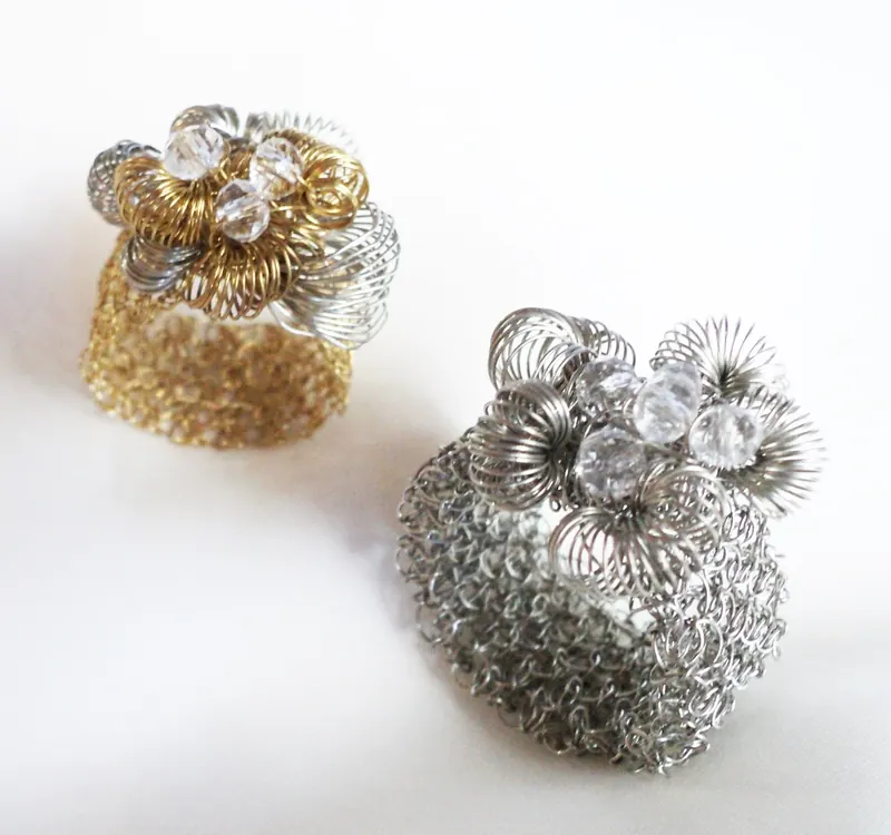 Dos anillos de malla tubular de alambre, uno plateado y otro dorado, con una roseta de rulos de alambre y cristales transparentes arriba