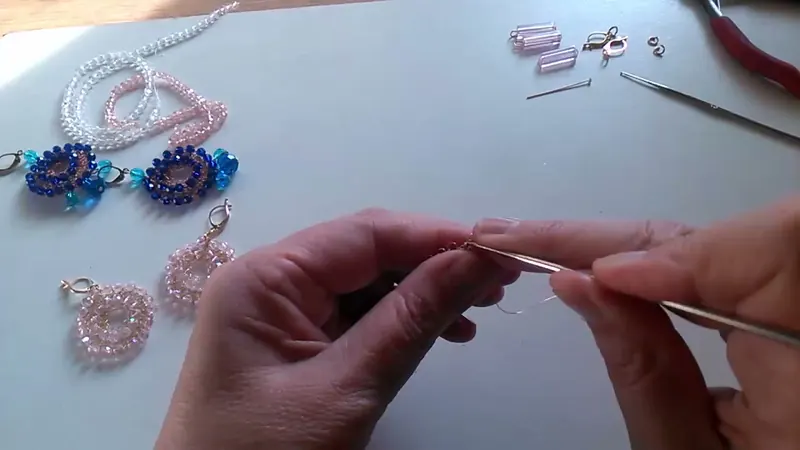 Las manos tejiendo la vuelta con la aguja de crochet mientras junto a la mesa hay otros aretes armados