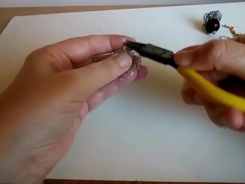 Una pinza de mango amarillo formando la anilla en el alambre, mientras la otra mano sostiene la perla forrada