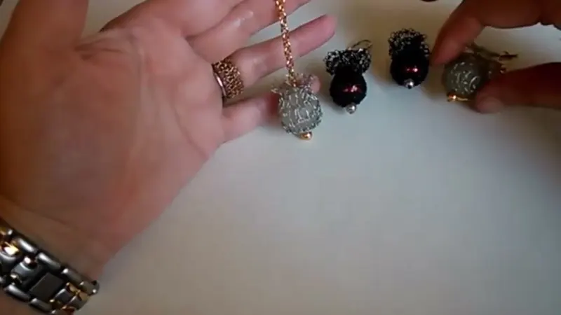 Una mano sostiene una cadenita con una perla forrada en malla de alambre gris; sobre la mesa hay otras tres perlas forradas, dos con malla negra y una con malla gris