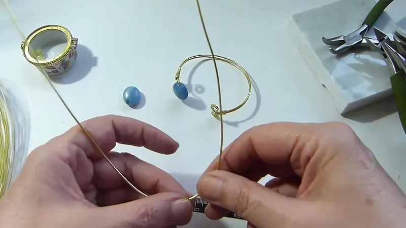 Manos que sostienen un tramo largo de alambre dorado, con un brazalete terminado y una gema azul de fondo