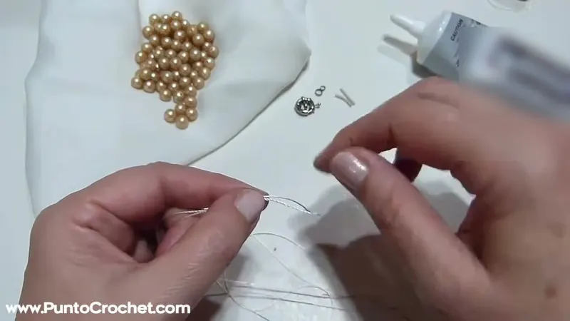 Dos manos sostienen un hilo fino sobre una mesa blanca donde hay perlas doradas sueltas, un cierre y anillas pequeñas