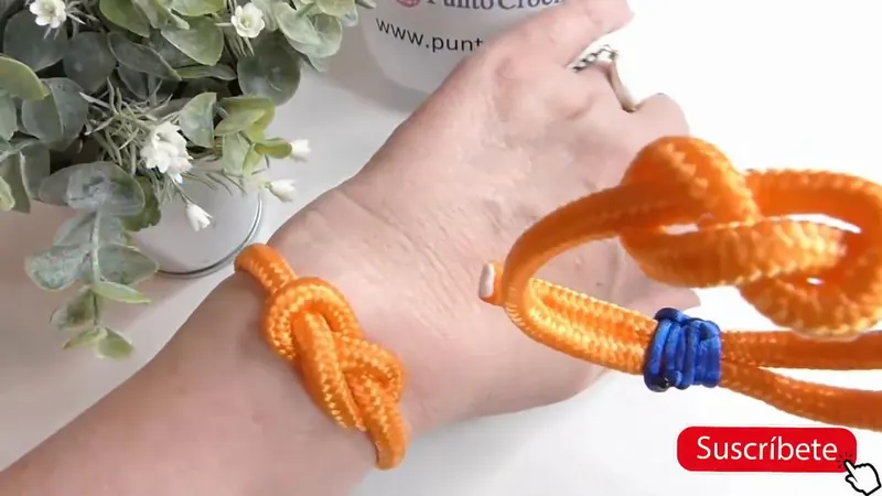 Brazalete de soga paracord naranja con nudo marinero y cierre de cordón azul, sobre una muñeca
