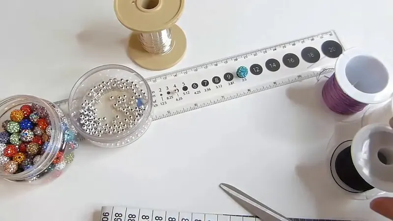 Mesa de trabajo con cuentas de colores, bolitas plateadas, una regla y carretes de elástico negro y de alambre delgado