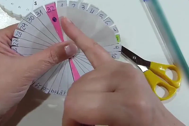 Dos manos sostienen un disco de papel y cartón con las ranuras numeradas del 1 al 32; a la derecha hay unas tijeras de mango amarillo y un bolígrafo