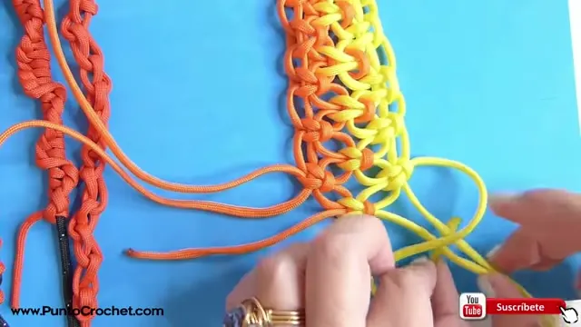 Tejido de nudos planos en naranja y amarillo con las manos formando un nudo con los cordones del centro
