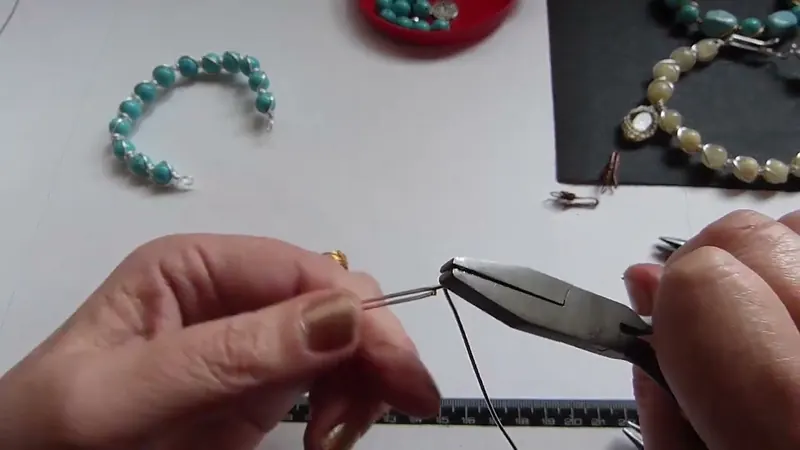 Las manos cerrando con una pinza plana el alambre doblado del cierre, con la pulsera armada de fondo