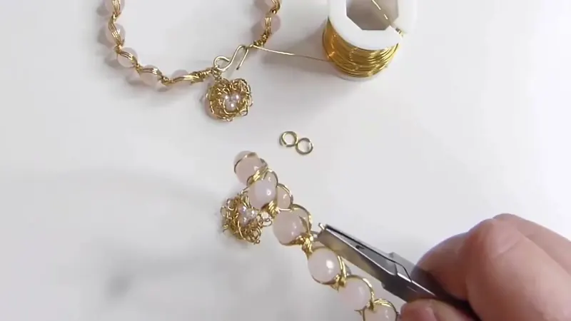 Una pinza cerrando la argolla que une el dije nido con la pulsera, con dos argollas sueltas sobre la mesa
