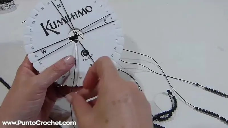 Disco kumihimo con hilos negros muy finos y una hebra de cristales negros a un lado, mientras una mano coloca un hilo