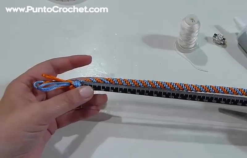La trenza redonda celeste y naranja tendida sobre una regla, con una mano que sujeta el extremo