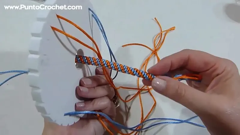 Dos manos sostienen la trenza redonda naranja y celeste ya larga, con los hilos sueltos aún sujetos al disco