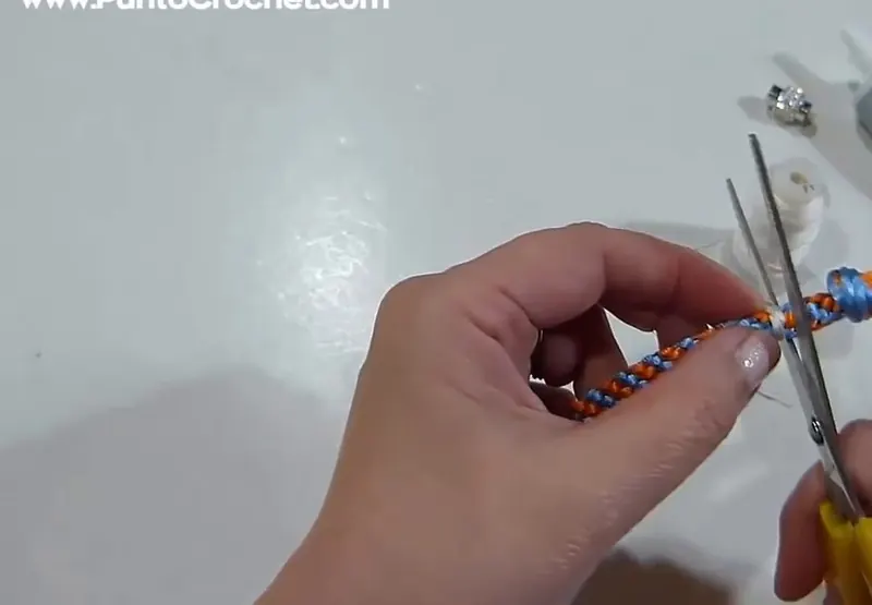 Una mano sujeta la trenza naranja y celeste con el extremo atado con hilo blanco mientras unas tijeras cortan los hilos sobrantes