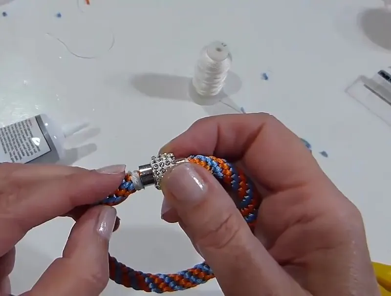 Dos manos colocan un capuchón magnético con pedrería en el extremo de la trenza naranja y celeste